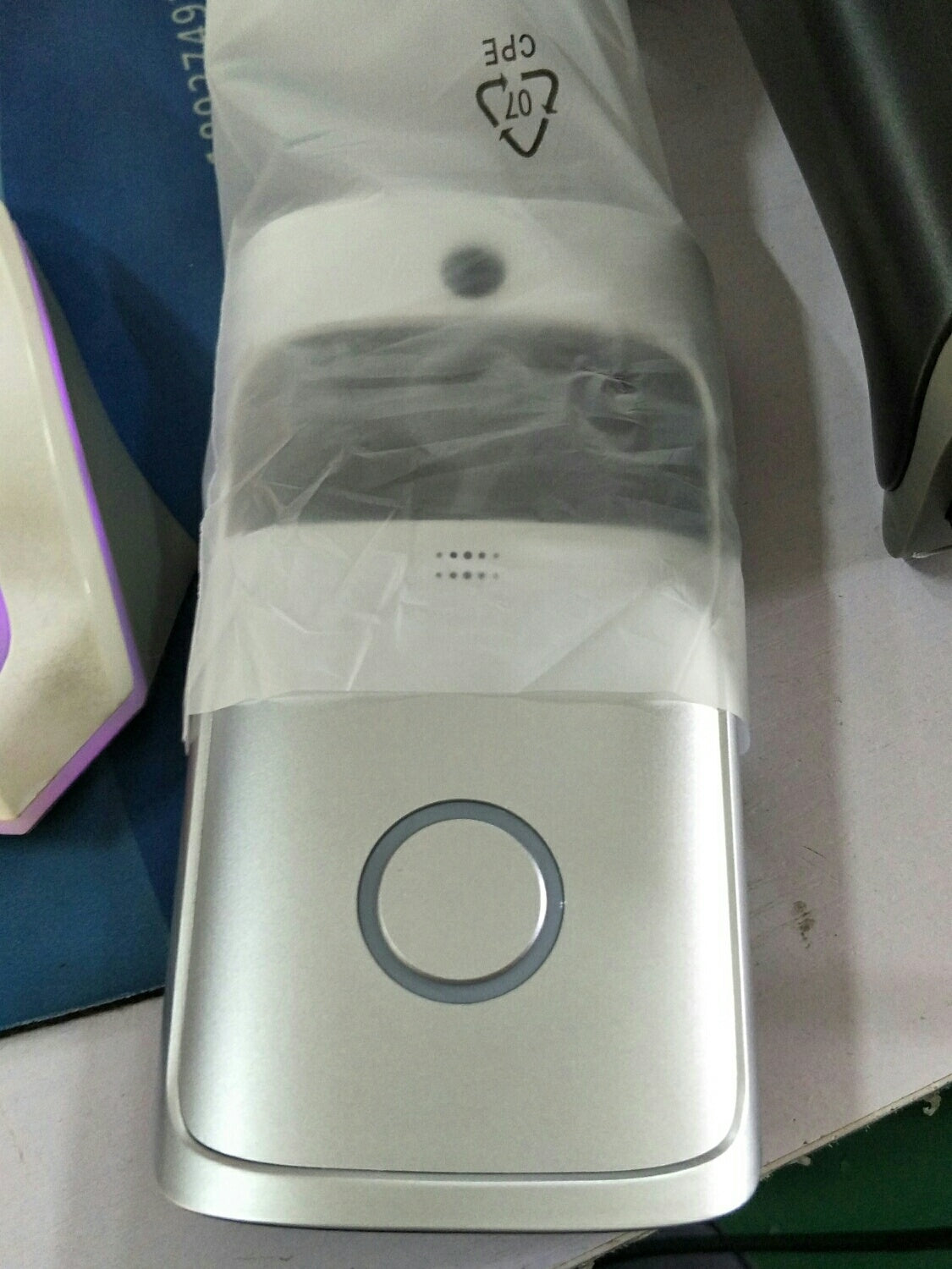 Silver smart doorbell with circular button and small camera lens above it wrapped in plastic packaging.