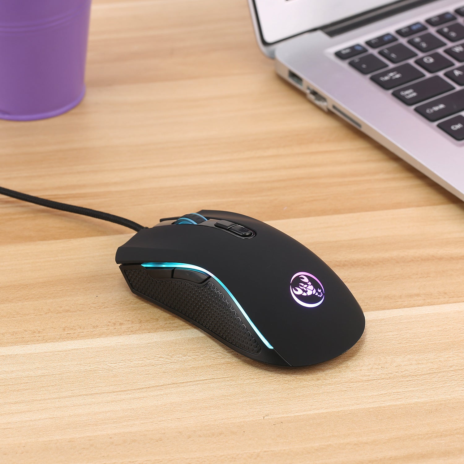 Black wired gaming mouse with RGB lighting and textured side grips on wooden desk next to laptop.